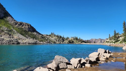A serene lake surrounded by rocky shores and lush green hills under a clear blue sky.