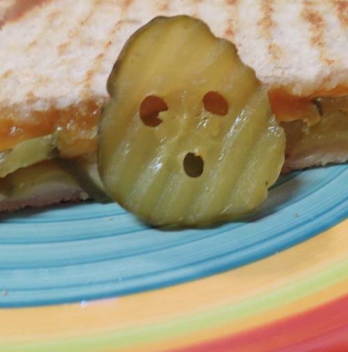 A close-up of a sandwich with a pickle slice shaped like a surprised face on a colorful plate.