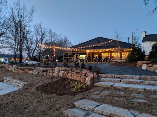 A cozy outdoor setting with string lights, a building, and stone pathways, surrounded by trees at dusk.