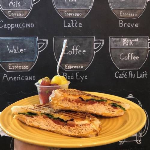 A hand holding a yellow plate with a grilled sandwich and a cup of fruit, against a chalkboard with coffee names.