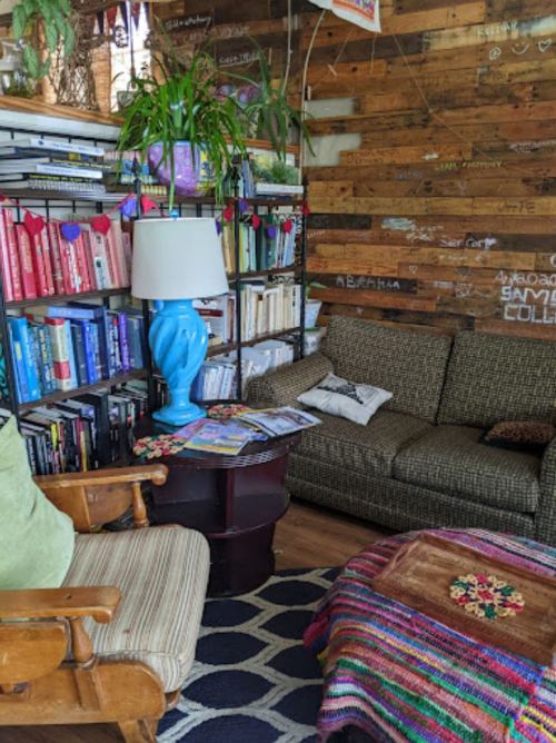 Cozy reading nook with a patterned rug, bookshelves, a blue lamp, and a green armchair in a rustic wooden setting.