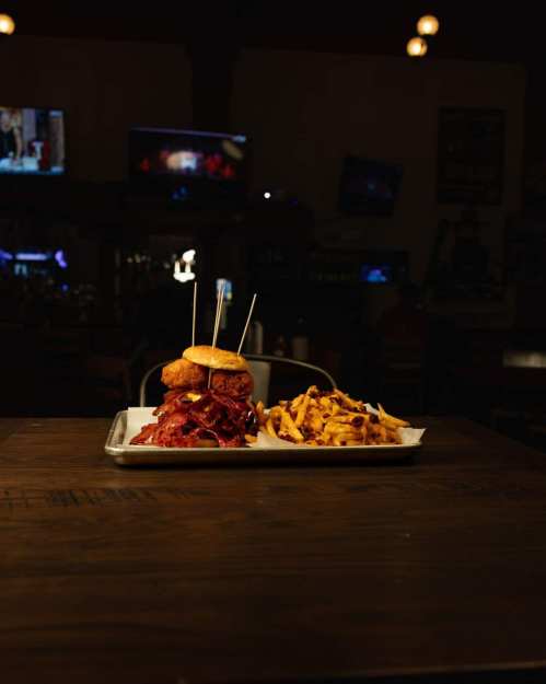 A plate with a stacked sandwich and loaded fries, set on a wooden table in a dimly lit restaurant.