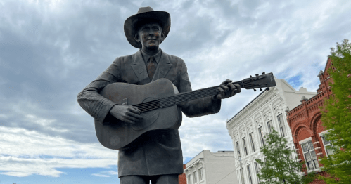 A bronze statue of a cowboy playing guitar, set against a cloudy sky and historic buildings.