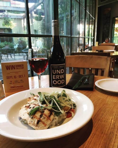 A plate of grilled chicken with vegetables, a glass of red wine, and a bottle of Pinot Noir on a wooden table.