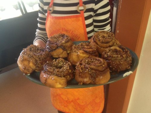 A person in an orange apron holds a platter of freshly baked cinnamon rolls topped with nuts and caramel.