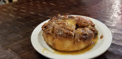 A cinnamon roll topped with nuts and drizzled in syrup, served on a small white plate on a wooden table.