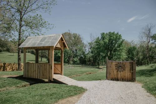A wooden gazebo with a metal roof beside a gravel path leading to a wooden gate in a green landscape.
