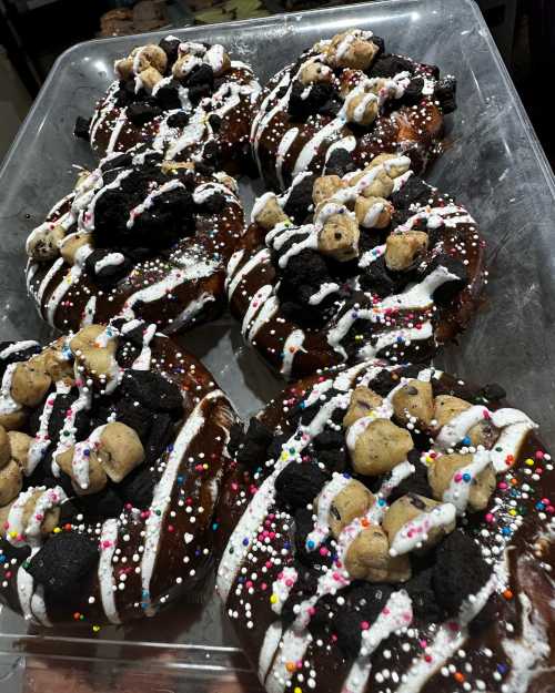 A tray of large, chocolate-covered donuts topped with cookie pieces, sprinkles, and white icing drizzle.