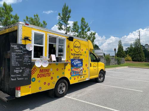 A bright yellow donut truck parked in a lot, featuring a menu board and colorful decorations. Trees and blue sky in the background.