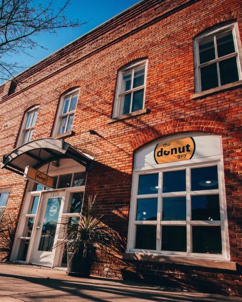Exterior of a brick building with large windows, featuring a sign that reads "the donut guy." Bright blue sky above.