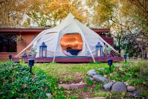 A cozy tent on a wooden deck surrounded by greenery and lanterns, set against a backdrop of autumn trees.