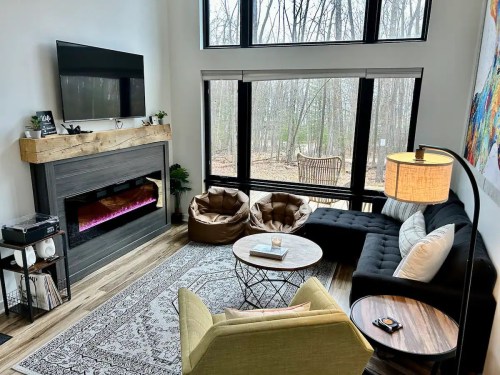 Cozy living room with large windows, modern furniture, an electric fireplace, and a view of trees outside.