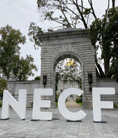Large white letters spelling "NECF" in front of a stone archway and trees, with a building in the background.