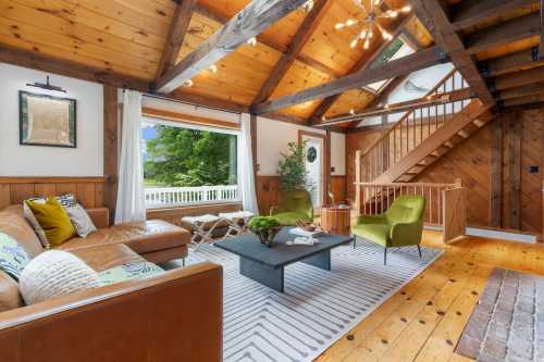 Cozy living room with wooden beams, a leather sofa, green chairs, and a staircase, featuring large windows and natural light.