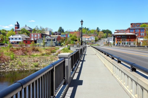 A scenic view of a riverside walkway with buildings and trees in the background under a clear blue sky.