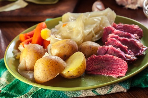 A green plate with boiled potatoes, carrots, cabbage, and slices of corned beef on a wooden table.