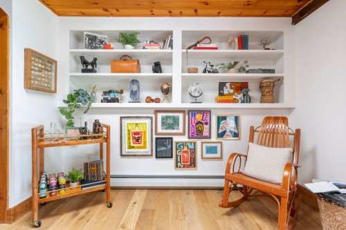 A cozy room featuring a wooden shelf with decor, a rocking chair, and a cart with plants and drinks.