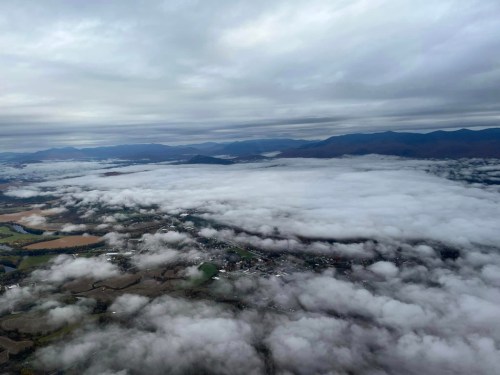 Aerial view of a landscape covered in thick clouds, with mountains in the background and patches of land visible.