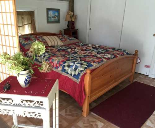 Cozy bedroom featuring a wooden bed with a colorful quilt, a small table, and a vase of flowers.