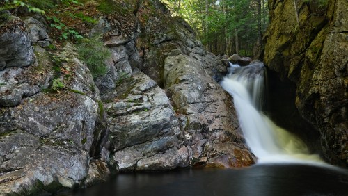 A serene waterfall cascades over rocky terrain, surrounded by lush green trees in a tranquil forest setting.