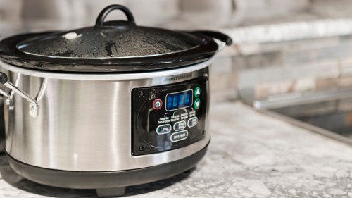 A stainless steel slow cooker with a digital display sits on a marble countertop in a kitchen.