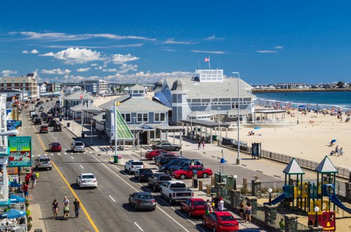 A sunny beach scene with a boardwalk, cars parked along the street, and people enjoying the sand and water.