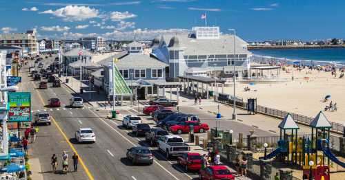 A coastal scene with a busy street, beach, and playground, featuring people, cars, and beachgoers under a blue sky.