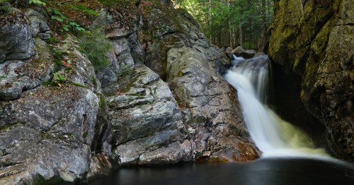 A serene waterfall cascades over rocky terrain, surrounded by lush green trees in a tranquil forest setting.