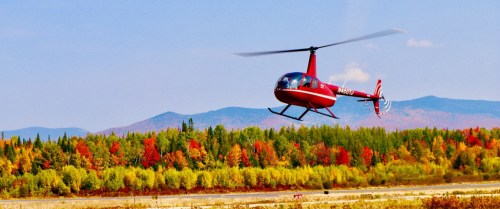 A red helicopter flies over a vibrant autumn landscape with colorful trees and mountains in the background.