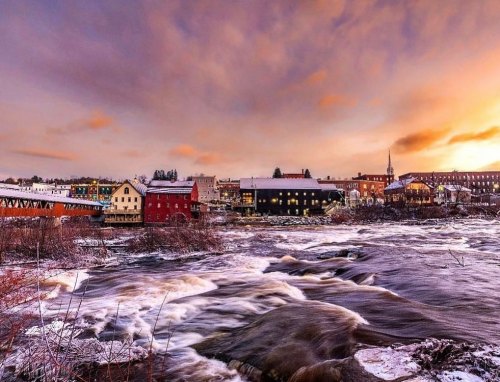 A scenic view of a river with flowing water, surrounded by colorful buildings and a sunset sky. Snow covers the ground.