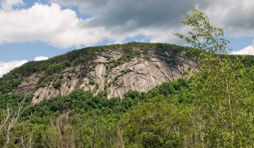 A rocky mountain face rises above lush green trees under a partly cloudy sky.