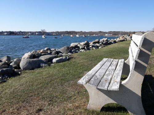 A concrete bench overlooks a calm bay with boats and a rocky shoreline under a clear blue sky.