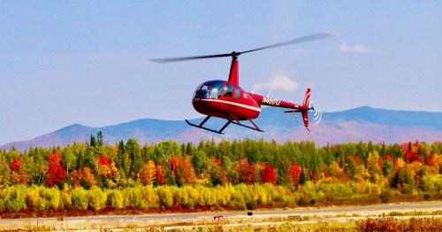 A red helicopter flies over a vibrant autumn landscape with colorful trees and mountains in the background.