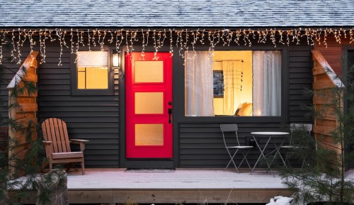 A cozy cabin with a bright red door, snow-covered porch, and festive lights hanging from the roof.