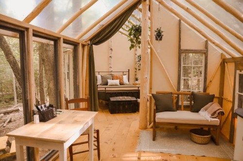 Cozy interior of a cabin with wooden furniture, a bed, and large windows overlooking a forested area.