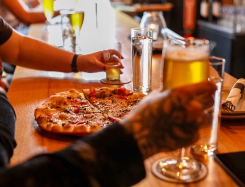 A close-up of a wooden bar with a pizza and drinks, including beer and water, being enjoyed by patrons.