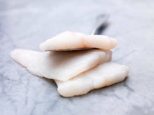 Three pieces of raw white fish fillet stacked on a marble surface, with a fork resting on top.