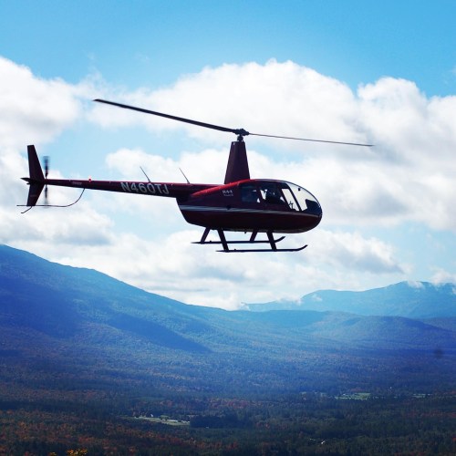 A red helicopter flying over a mountainous landscape with blue skies and scattered clouds.