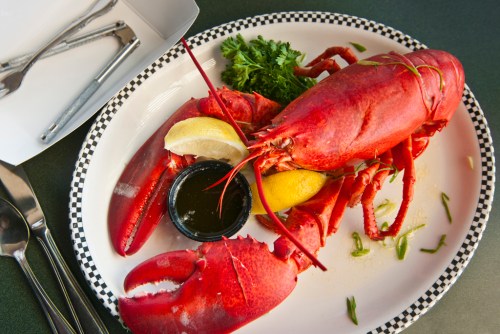 A cooked lobster on a plate with lemon, parsley, and a small dish of dipping sauce, alongside utensils.