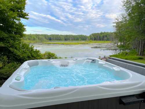 A hot tub with bubbling water overlooks a serene lake surrounded by lush greenery and a cloudy sky.