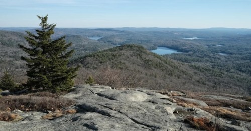 A scenic view from a rocky mountain top, overlooking lush valleys and lakes under a clear blue sky.