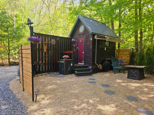 A cozy tiny house with a green roof, surrounded by trees and gravel, featuring a small patio and decorative elements.