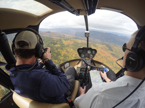Two pilots in a helicopter cockpit, one taking photos while the other navigates with a map, surrounded by autumn scenery.