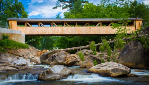 A wooden covered bridge spans a rocky river, surrounded by lush greenery and a blue sky with clouds.