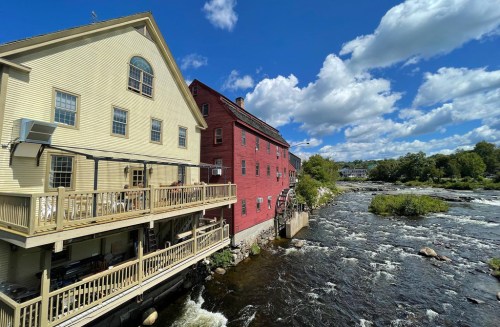 A scenic view of two buildings by a river, with a wooden deck and lush greenery under a blue sky with clouds.