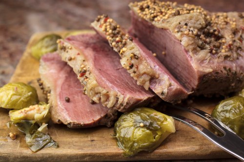 Sliced roast meat with a mustard crust, served with Brussels sprouts on a wooden cutting board.