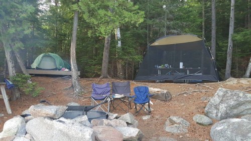 A campsite with two tents, camping chairs, and a fire pit surrounded by trees and rocks.