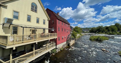 A scenic view of two buildings by a river, one yellow and one red, with a blue sky and fluffy clouds above.