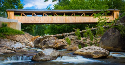 A wooden covered bridge spans a rocky river, surrounded by lush greenery and a clear blue sky.