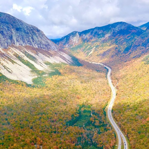 A winding road through a vibrant valley surrounded by mountains and colorful autumn foliage under a cloudy sky.